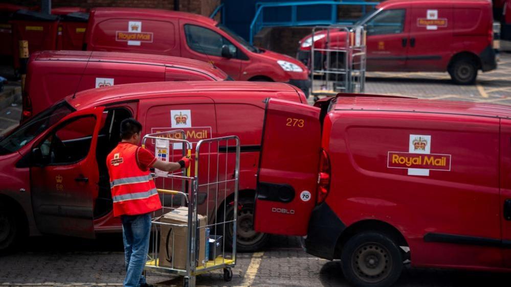 Royal Mail worker loading parcels onto a Royal Mail van. There are five red Royal Mail vans in the image. 