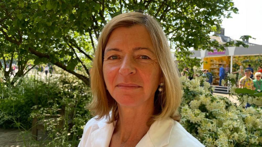 Laura Lee with medium-length blonde hair and white jacket in a garden