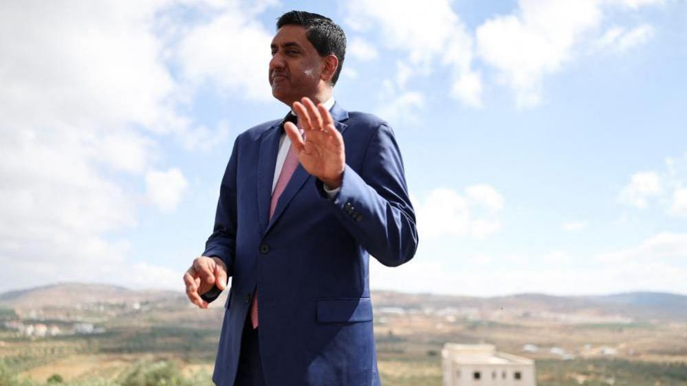 Khanna, who is dressed in a blue suit, white shirt and pink tie, raises his hand as he is photographed outdoors with a rural town landscape behind him
