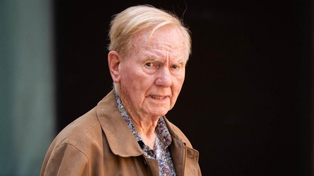 An older man in a pale brown jacket and paisley shirt looks toward the camera with a serious expression.