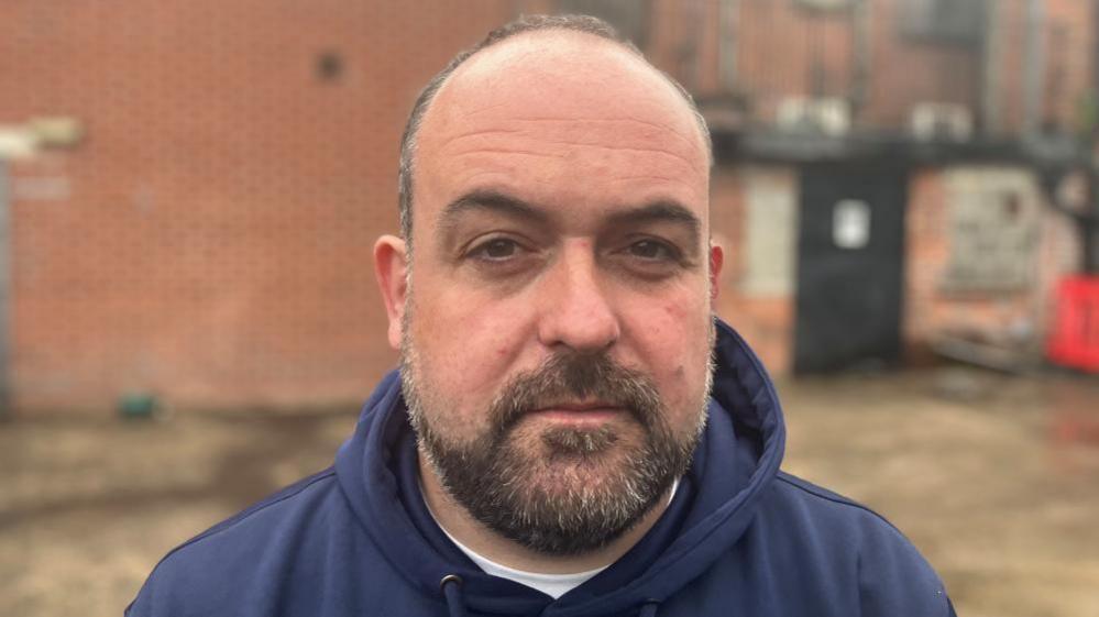 Paul McCusker is looking directly at the camera. It is a head and shoulders shot. He is wearing a navy hoodie and has short brown hair with a beard. Behind him red brick walls can be seen.