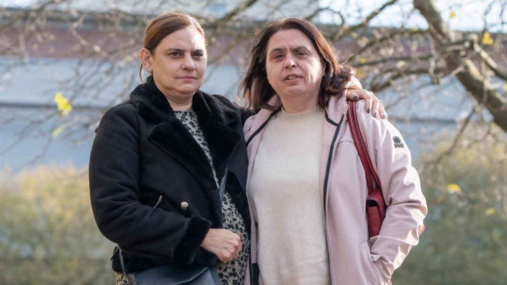 Two women stood outside Bradford Crown Court. One wearing a black jacket has her arm around the other, dressed in a pink jacket and top