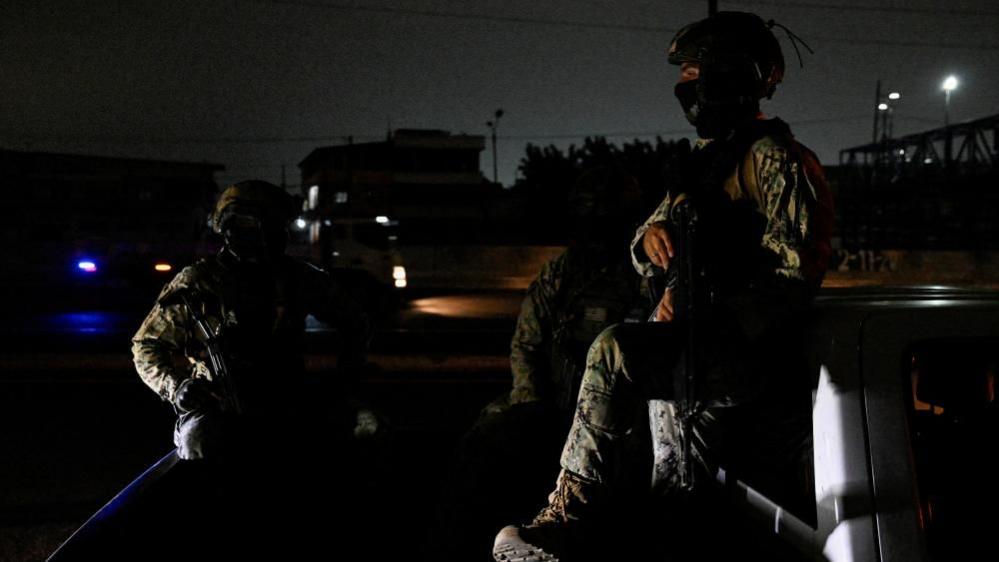 Two people in military fatigues sit in the bed of a truck in a street enveloped in darkness