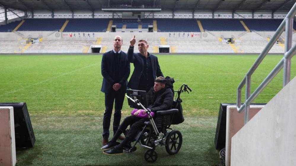 Prince William stands alongside Kevin Sinfield and Rob Burrow during a visit to Headingley Stadium in Leeds. Kevin Sinfield is pointing upwards as they stand at the side of the pitch.