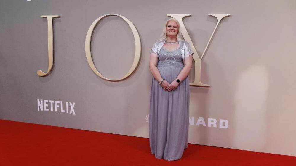 Louise Brown is wearing a long grey dress with a silver short sleeved jacket. She has long blonde hair and is smiling while standing in front of a promotional board for the film, 'Joy' at a film festival. The board also mentions Netflix.