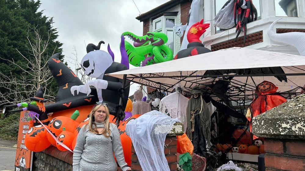 An end of terrace house barely visible underneath lots of halloween decorations, including dozens of huge inflatable ones. Smaller decorations are all over the front garden for people to see walking into the garden path.
Claire Cox stands in front wearing a grey jumper, dwarfed by the size of the decorations.