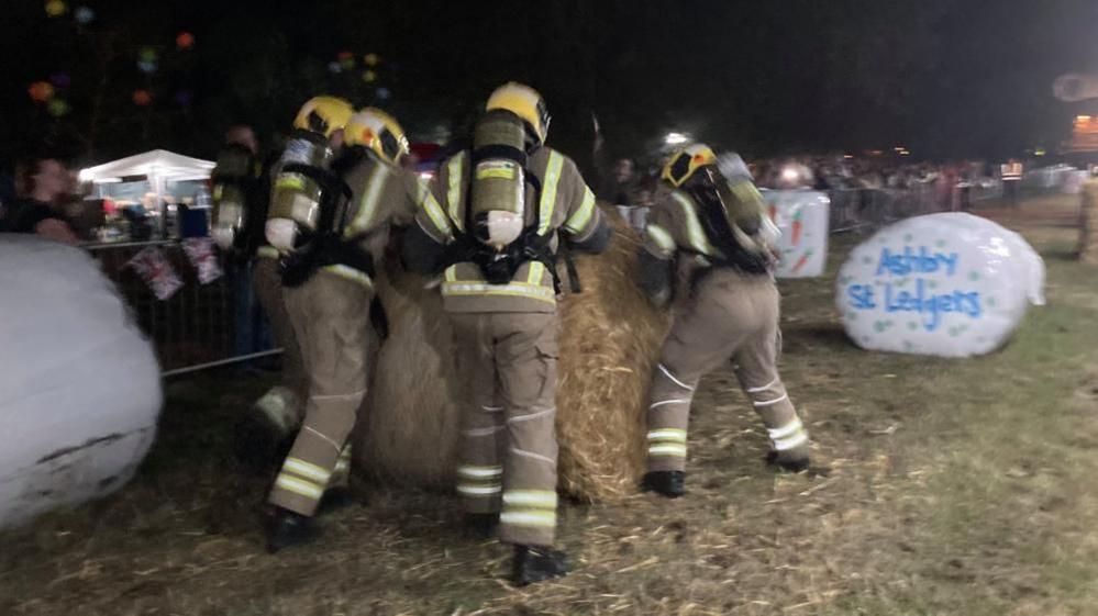 Four firefighters wearing beige firefighting clothes, yellow helmet and tank push a large hay bale around a grass course.  A static bale with a white cover bears the legend "Ashby St Ledgers".