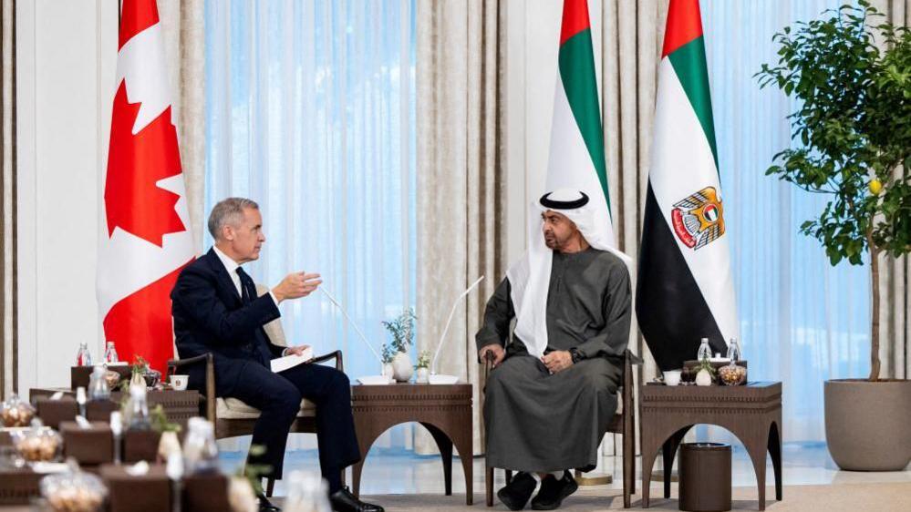 Sheikh Mohamed bin Zayed Al Nahyan, President of the United Arab Emirates, meets with Canadian Prime Minister Mark Carney at Al Shati Palace in Abu Dhabi, United Arab Emirates, November 20, 2025. The two are seated across from each other. Carney is wearing a black suit, while the Al Nahyan is wearing a traditional Emirati thobe and white gutra. Behind them are the UAE and Canadian flags. 