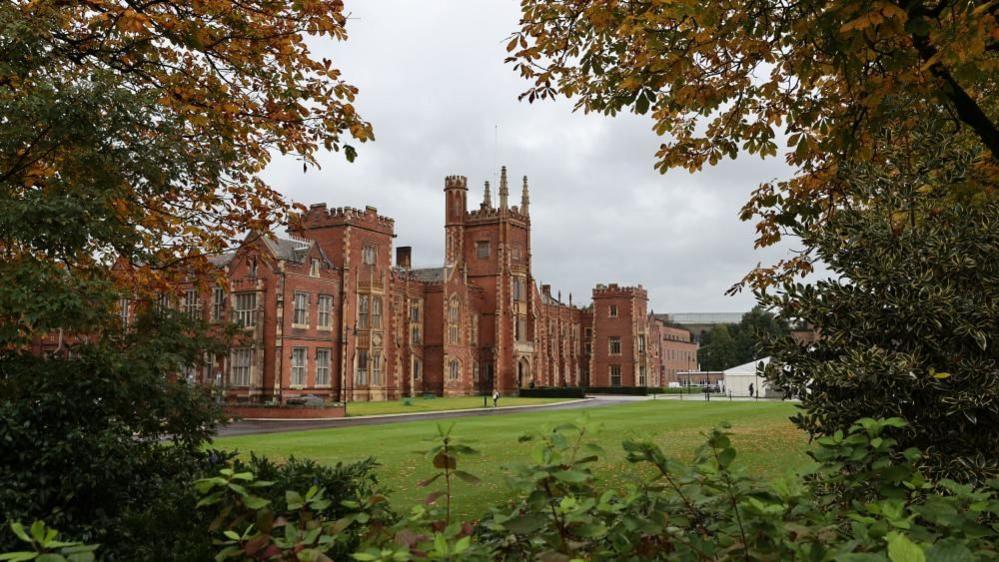 A general view of the main building of Queen's university - a red brick grand building. There are leafy green trees in the foreground