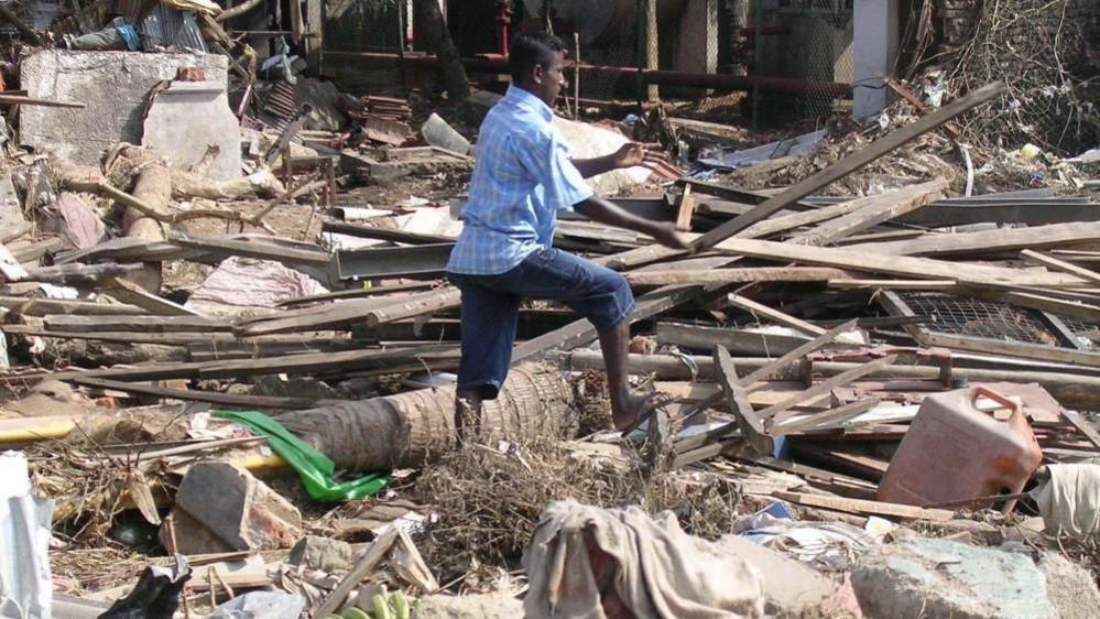 A man wearing blue jeans and a shirt stands in the middle of a pile of debris from wrecked buildings