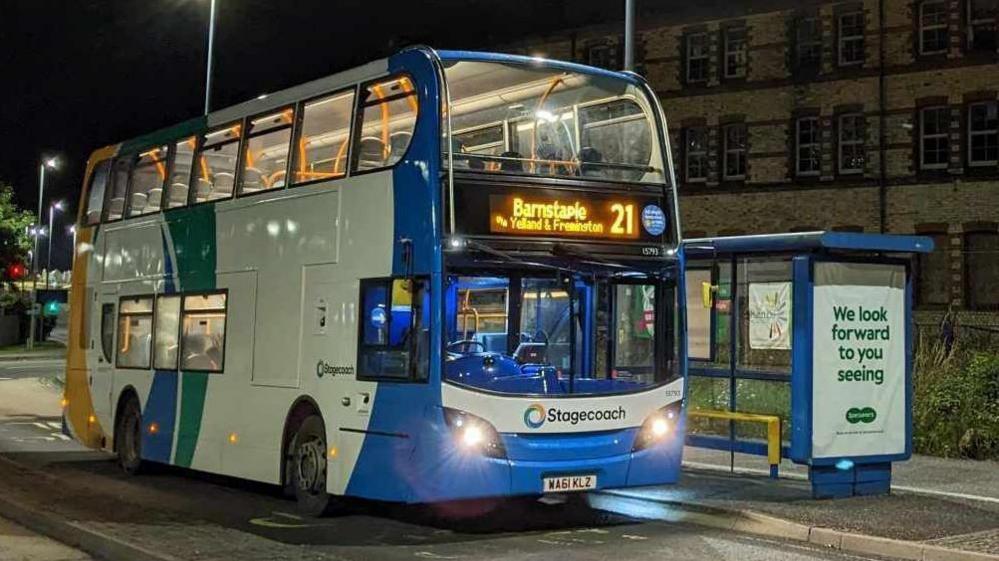 A Stagecoach bus which has been painted primarily white with a light blue front with yellow, green and light blue stripes at the back. It is parked at a blue bus stop at night.