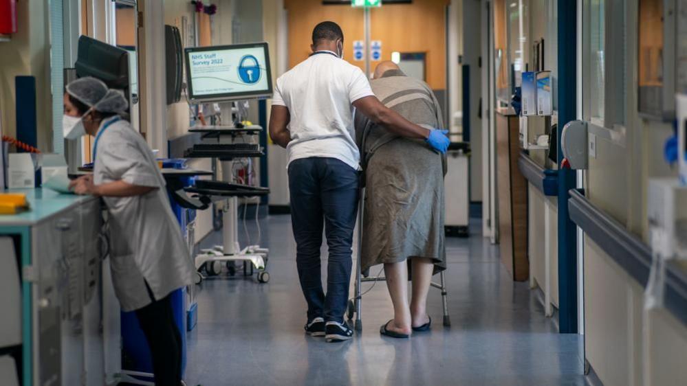 Seen from behind, a male nurse escorts an elderly man with a walker down a hospital corridor