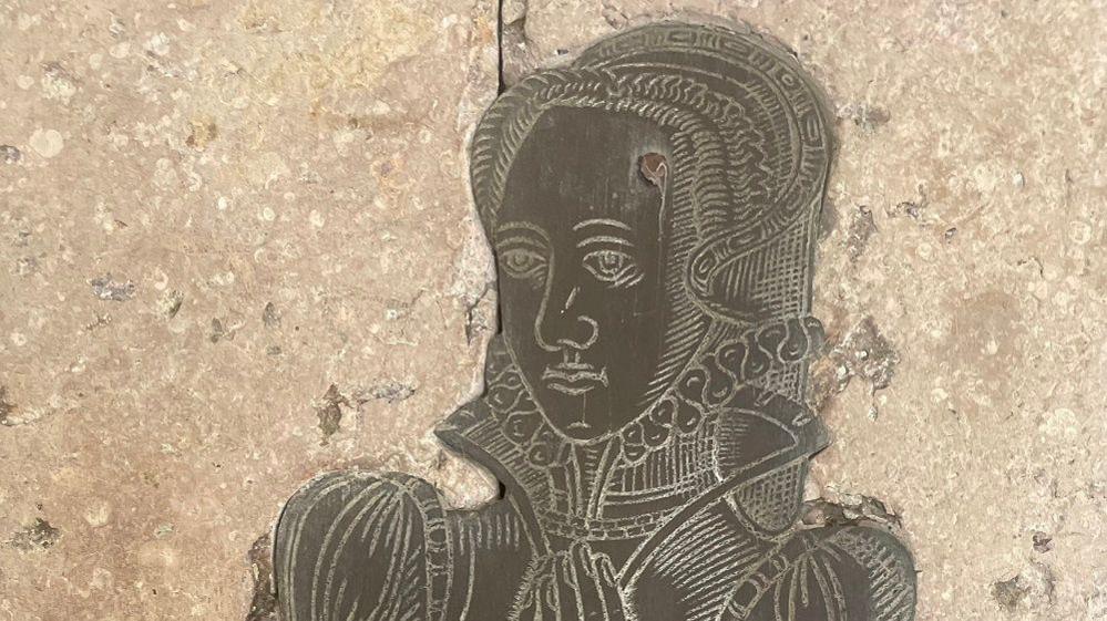 Part of an old brass etching of a woman in Tudor dress attached to a stone wall.