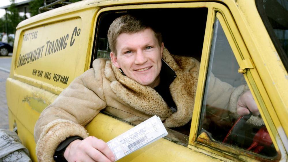 Ricky Hatton, wearing a sheepskin jacket, sits in the driver's seat of a little yellow van. it has a sticker on it that says 'Trotters Indpendent Trading Co' on it.