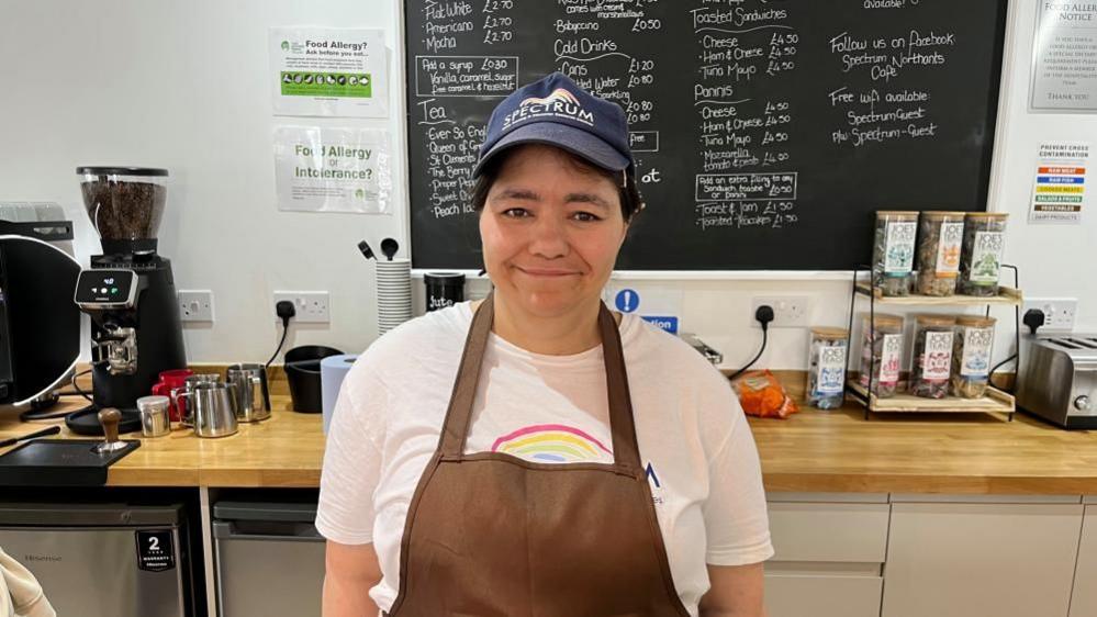 A woman in a T-shirt, cap and an apron standing in front of a coffee machine and counter