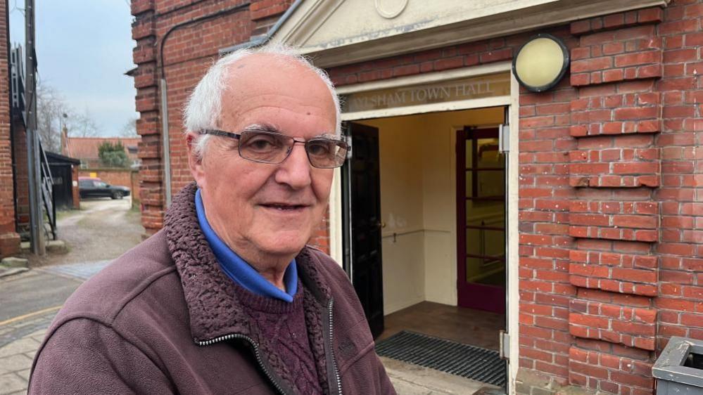 Brian Schuil is wearing a burgundy fleece with a burgundy knitted jersey and blue collared polo shirt beneath. He has receding white hair, and is wearing glasses. He is pictured outside the entrance of Aylsham Town Hall, a Georgian building with decorative red brickwork.