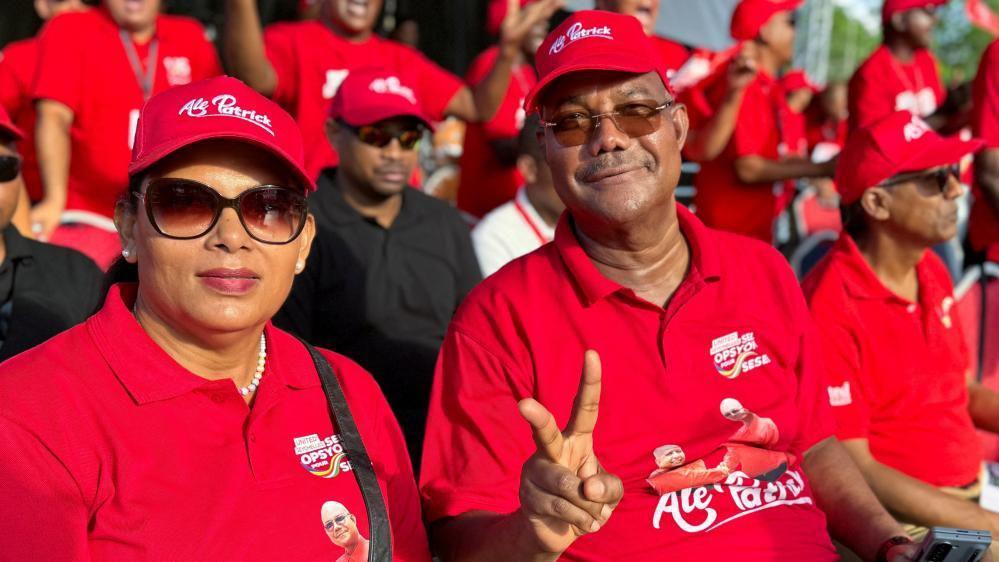 Dressed in a red T-shirt and cap, Patrick Herminie, flanked by his wife Veronique, flashes a two finger salute on September 27, in Victoria