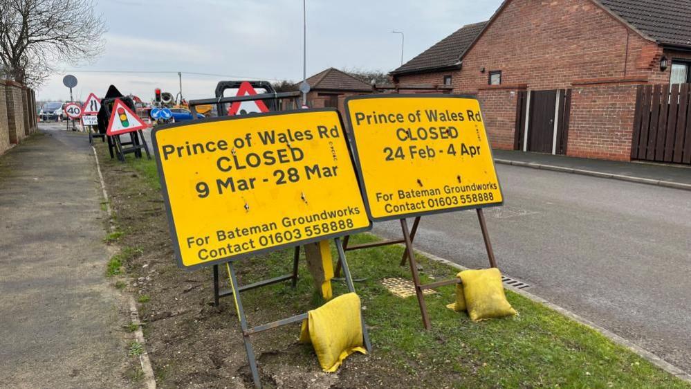 Yellow road signs that state Prince of Wales Road in Caister being closed to traffic at the junction of Jack Chase Way from 24 February to 4 April. It states For Bateman Groundworks contact 01603 558888. The images shows part of Prince of Wales Road and Jack Chase Way intersecting at the junction. There is a footpath and grass verge in the foreground and a brick wall in the right of the image.