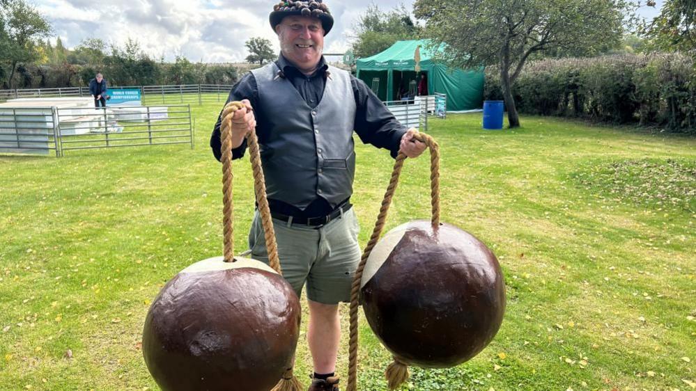 A man, wearing shorts, a black leather waistcoat, black shit, black hat, with conkers round the rim, and holding two giant pretend conkers on rope. She is smiling and looking at the camera. He is standing on green grass.