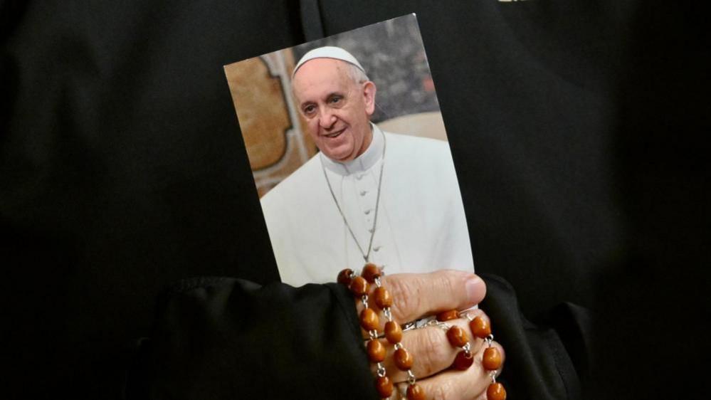 In a close-up image of her hands, a nun clutches an image of the Pope and rosary beads