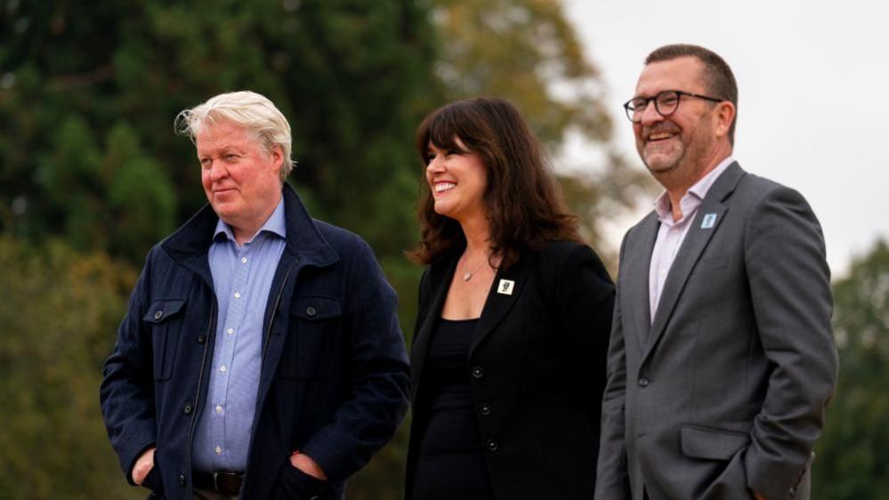 Earl Spencer with short white hair, a blue jacket and blue shirt, with his hands in his pockets, Caroline Redman Lusher with long dark hair and a black top and jacket, and Nigel Varndell with short dark hair and beard, wearing a grey suit with his hands in his pockets. There are trees behind them.