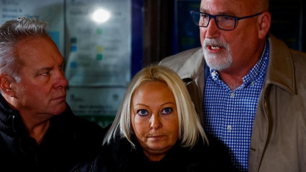 Radd Seiger with short grey hair and beard, wearing glasses, a grey jacket and blue and white shirt. He is standing behind Charlotte Charles, who has long blonde hair and is wearing a dark coat, and Bruce Charles, who has medium-length grey hair and is wearing a dark coat.