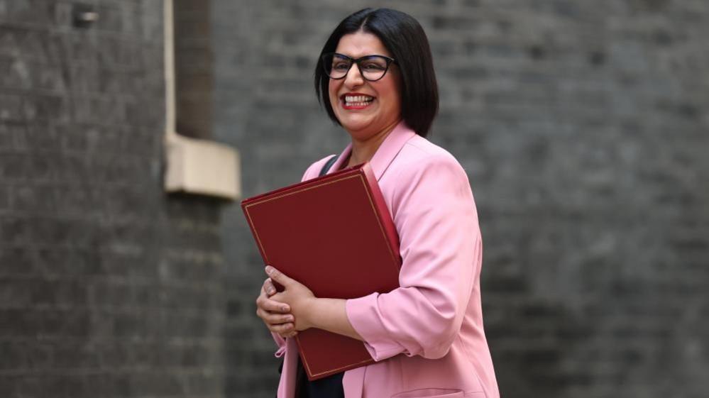 Shabana has a wide smile as she wears a pink blazer and holds a red binder. she's walking past a grey brick wall.