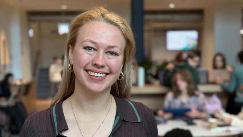 Wearing a brown collared top with a light blue trim, Julia smiles opened mouthed at the camera. She wears gold hooped earrings with her blonde hair loose down her back. 