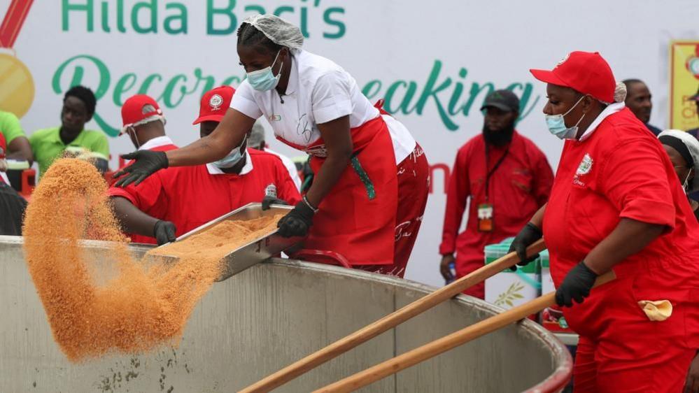 Hilda Baci pours chili powder into a giant pot. She is wearing a white chef's top and red apron. Helpers- all dressed in red - are standing around the pot.