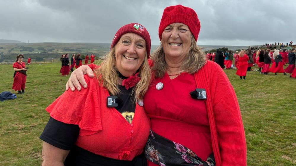 Two women wearing red dresses and red beanie hats. In the background is a group of people wearing red dresses in a field.