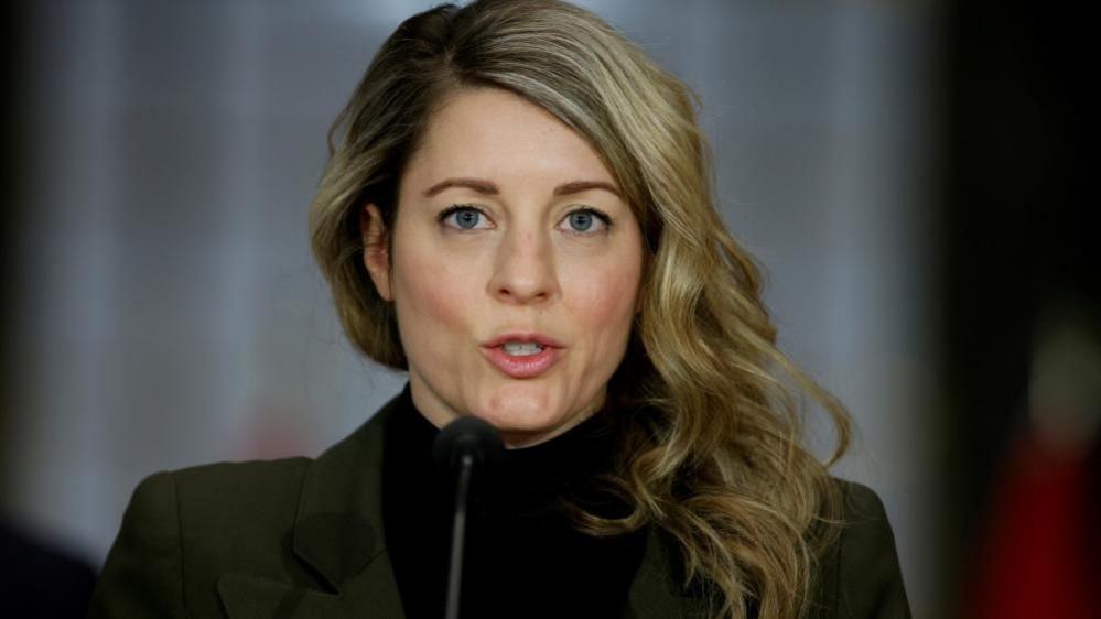 Canada's Minister of Foreign Affairs Melanie Joly at a news conference, stands at a microphone wearing a dark suit coat and a black turtleneck sweater