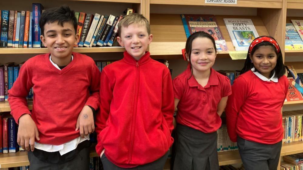 A mixed gender group of Fishergate Primary School pupils stand in front of wooden library bookshelves. They wear a mix of school uniforms; red jumpers, polo shirts and fleeces, with grey trousers.