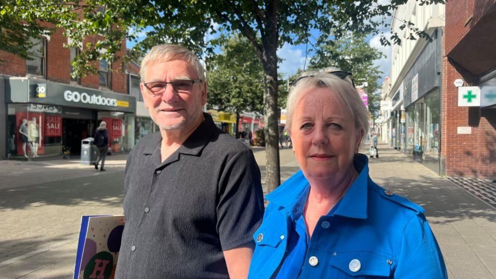 Debbie Holmes is wearing a blue jacket, and has blonde hair, with her sunglasses perched in her hair. Her husband is standing to her right, and he is wearing a black shirt, and is holding a large, flat, paper bag. They are standing on London Road North, with shops lining either side of the street, with some trees behind them.