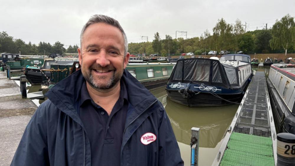 Chris Barlow with short dark hair and a beard wearing a blue polo shirt and blue raincoat with red Whilton Marina logo. There are narrowboats in mooring positions at the marina behind him, and trees beyond them.