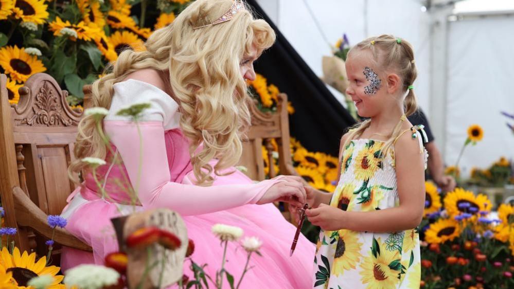 A woman dressed as a Disney princess in a pink dress and blond wig holds the hand of a young girls. They are smiling at each other and there are sunflowers in the background. 