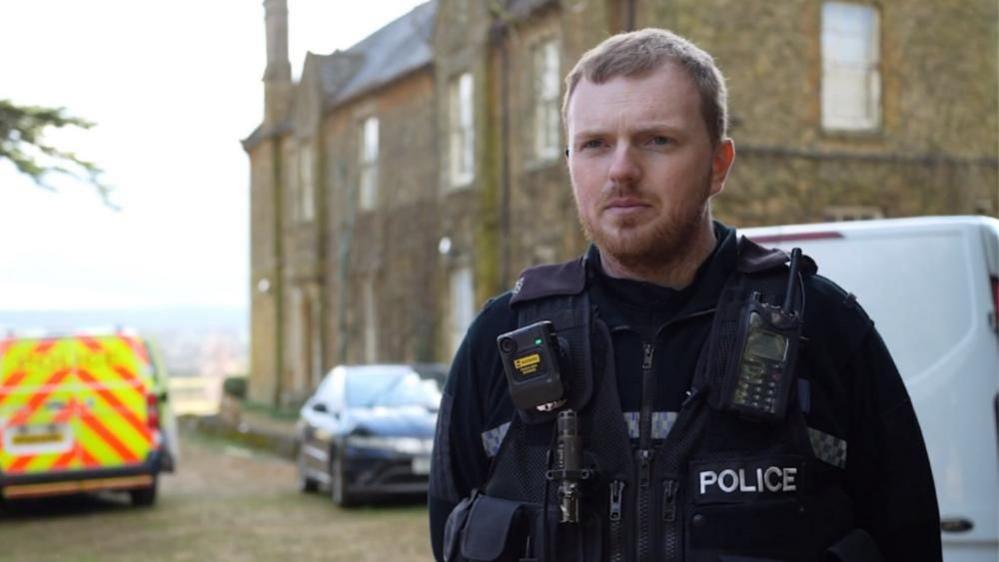 PC Partridge with short brown hair and beard wearing a black police uniform with radios attached below the shoulder. There is a yellow and red police fan behind him, with a car and a white van. There is a stone-building two-storey house in the background.