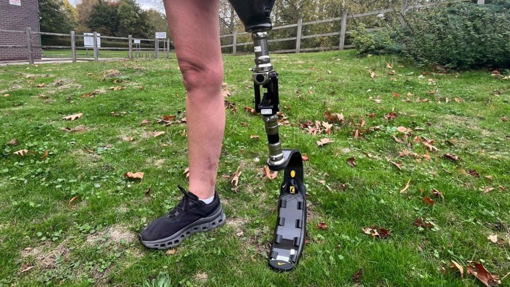 A close up of Lisa's prosthetic blade. It's made of black plastic and metal, and is about the size of her right leg. The lower third of it is a curved, flexible structure that allows her to push off and run. She's stood on grass, with a wooden fence in the background.