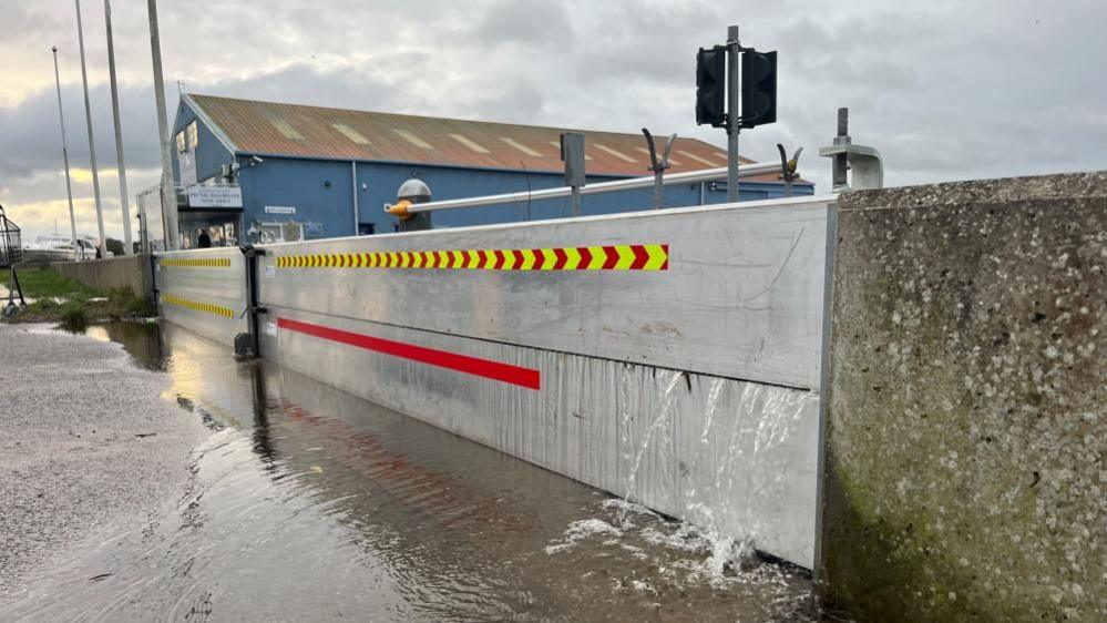 The flood gate at Potter Heigham is made of aluminium and is set into the concrete flood wall. A trickle of water is seeping through it and there is a puddle on the ground. Behind the gate is a car park barrier, and beyond that a blue boat shed belonging to Herbert Woods boat yard.