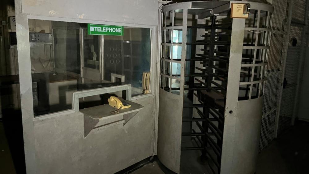 A metal grey-painted turnstile gate and security office. A telephone is mounted on a shelf, beneath a window to the security office. A green sticker stating: "telephone" is stuck onto the glass.