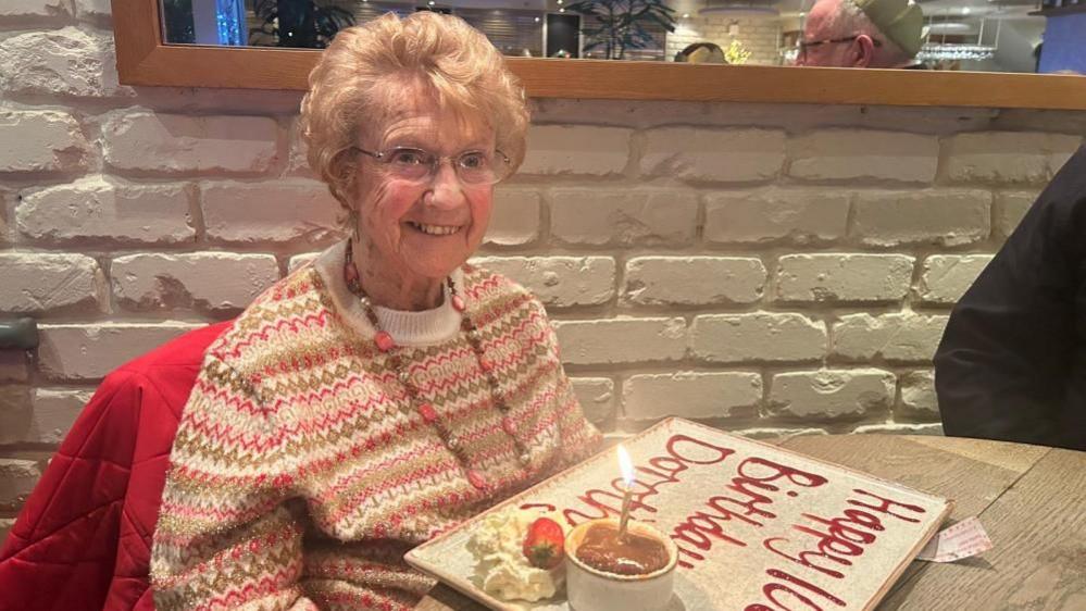 An old woman wearing a pink jumper sits in front of a big plate which says 'Happy Birthday Dorothy' in icing. 