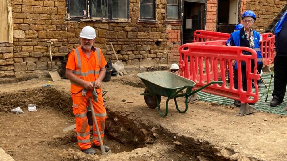 Man in orange hi-vis digs with a pickaxe on an archaeological site