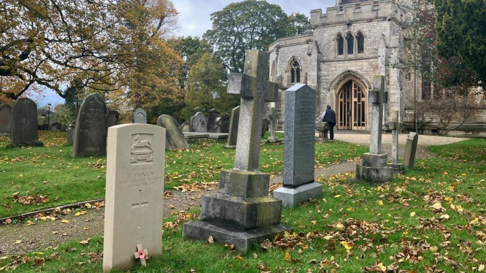 Emmanuel J. Foxton's Commonwealth War Grave in St Helen's churchyard. The grave is a light-coloured stone, bearing an engraving of a horse above a banner, Emmanuel J. Foxton's name, date of death and a cross. It is positioned next to an older stone cross headstone, in a row with other grey headstones and on a grassy area divided by a path. The church, a light grey stone building, can be seen in the background of the image.
