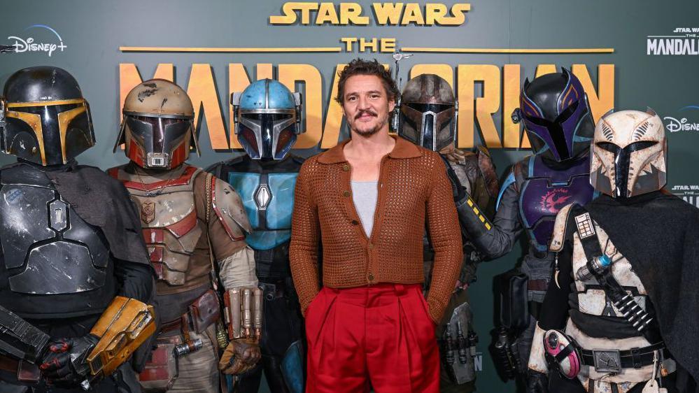 Pedro Pascal stands with his hands in his pockets in front of six costumed characters from The Mandalorian.