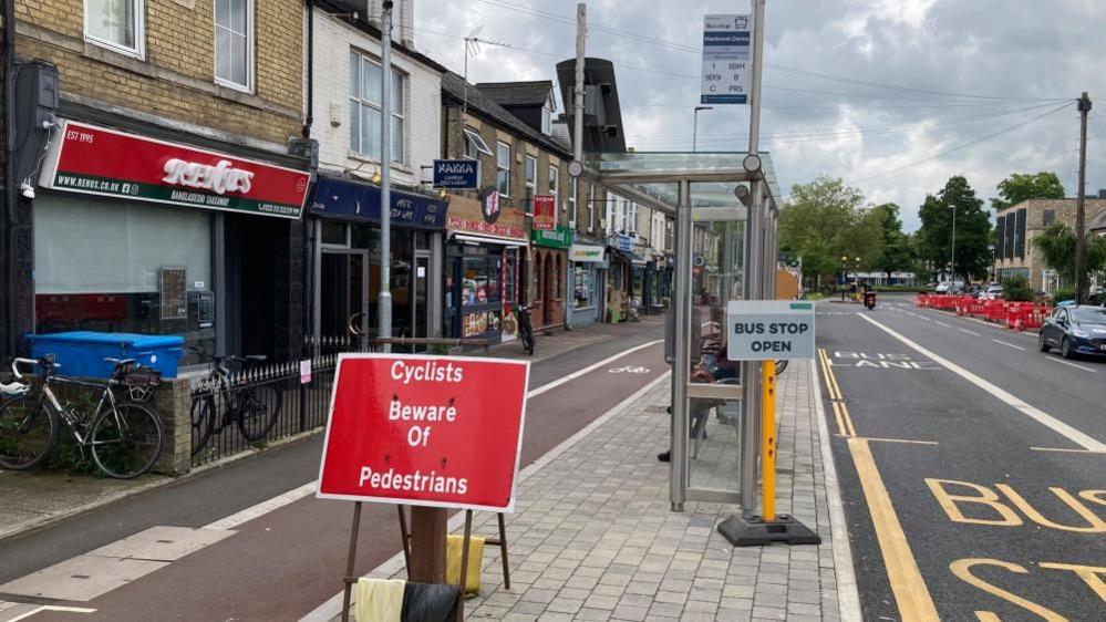 A bus stop next to the road on a street, with shops lining the pavement to the left of the bus stop. There is a cycle lane indicated in dark red tarmac between the bus stop and the pavement, with a red sign reading "Cyclists beware of pedestrians" in front of the bus stop. There is a glass bus shelter and sign.