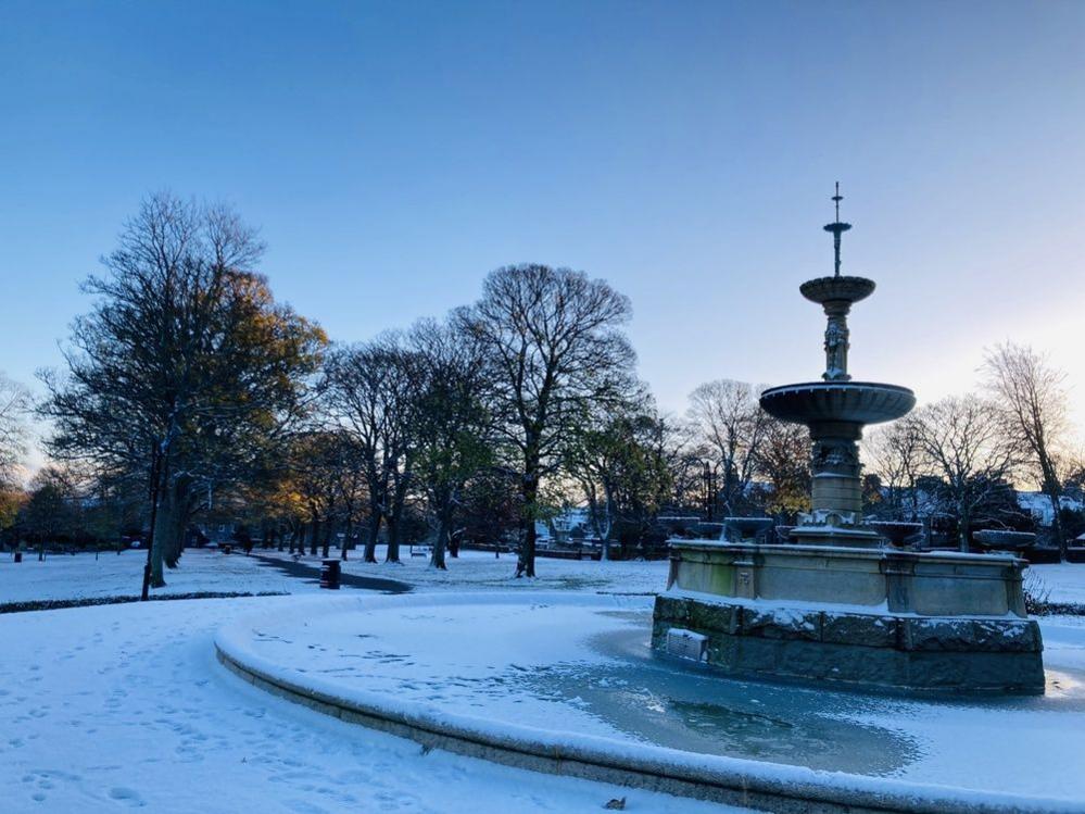 A park is covered in snow, with bare trees lining a path towards a fountain, which is still and covered in snow with sections of ice. 