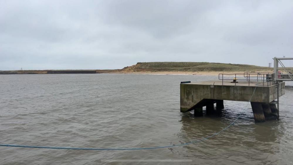 A concrete "dolphin" - a structure used to moor ships alongside while they load or discharge cargo, usually stone or sand minerals. In the far distance is the undeveloped southern terminal, which is made of sand, much of which is covered in sand. In the foreground is sea water in the outer harbour.