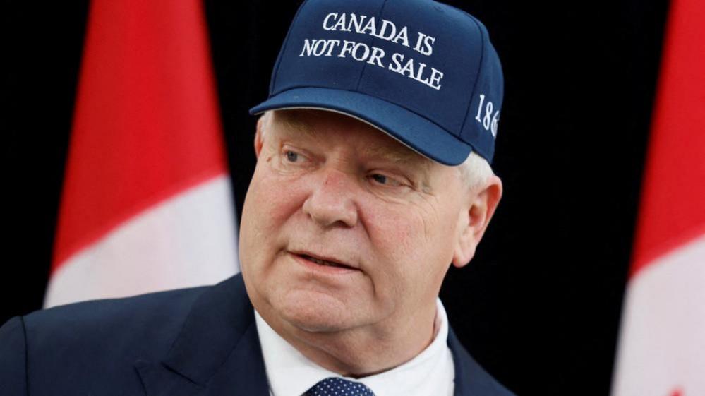 Ontario Premier Doug Ford is wearing a dark blue baseball cap with the logo 'Canada is not for sale' across the front. He is standing before two Canadian flags in the background as he looks to the right. 