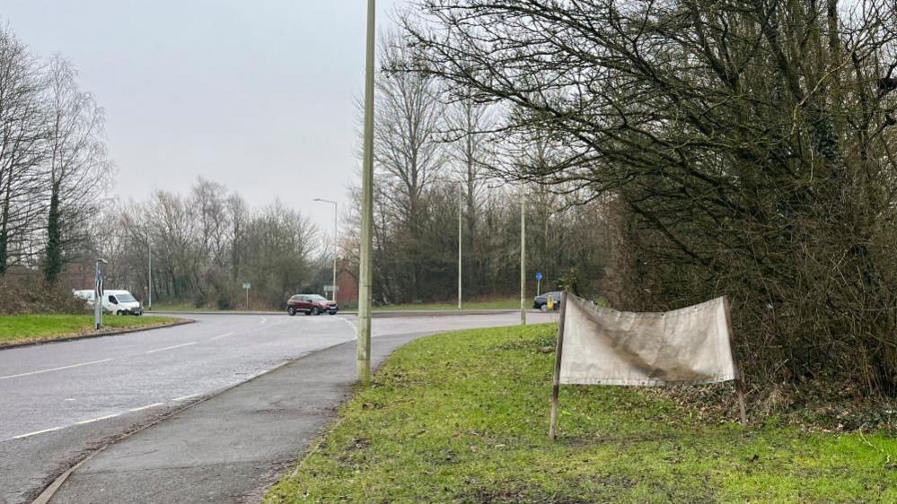 Picture of the back of the sign on a grassy verge with pavement running alongside. The sign is white, and held up by two wooden poles. Part of the roundabout is visible, with three cars in the distance. There are trees and shrubbery along the edges of the grassy areas. 