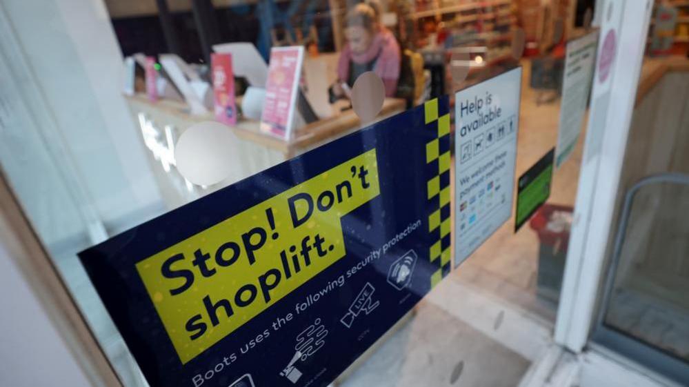 A sign in a shop window says 'Stop! Don't shoplift. Boots uses the following security protection'. A blurred out customer can be seen using a self service till inside the store.
