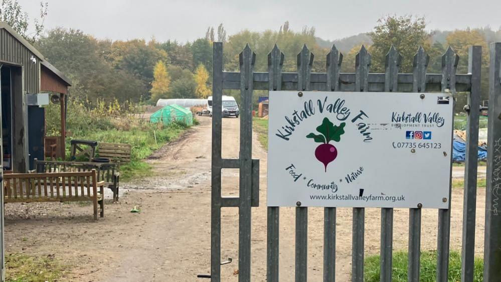 A sign on a metal gate reads: Kirkstall Valley Farm, Food, Community, Nature. Behind it is a farm shed, two benches and plots of allotment land